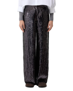 Someone stands against a plain background wearing the TWP Adieu for Now Sequin Pant in black, paired with a white shirt, gray sleeves, and dark shoes.
