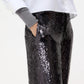 A person wearing a white button-up shirt, gray sweater sleeves, and TWP's Adieu for Now Sequin Pant in black with one hand in their pocket.