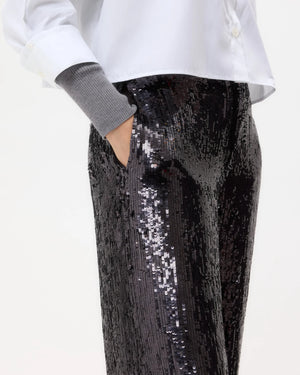 A person wearing a white button-up shirt, gray sweater sleeves, and TWP's Adieu for Now Sequin Pant in black with one hand in their pocket.