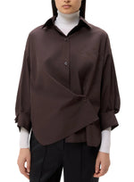 A woman with straight, dark hair wears the TWP New Earl Shirt in a relaxed, oversized fit over a white turtleneck and black pants, standing against a plain white background.