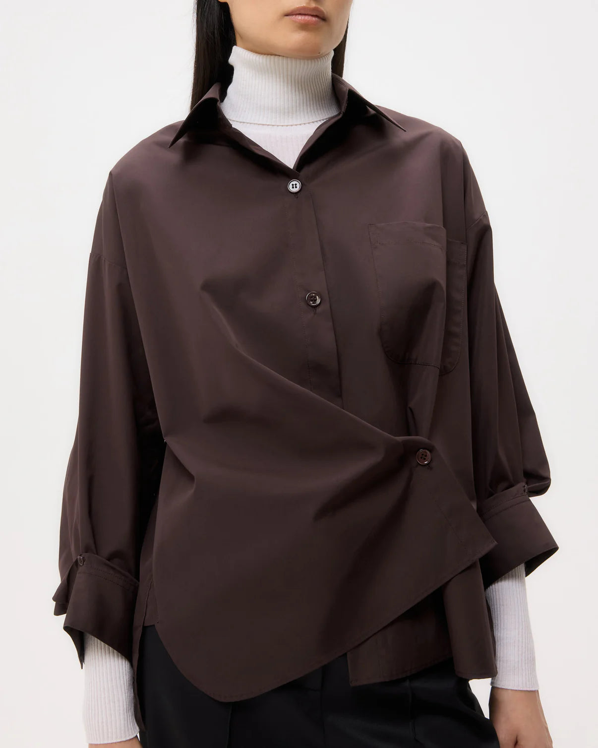 A person wears the TWP New Earl Shirt in dark brown, styled asymmetrically over a white turtleneck, pictured from shoulders to hips against a plain background.