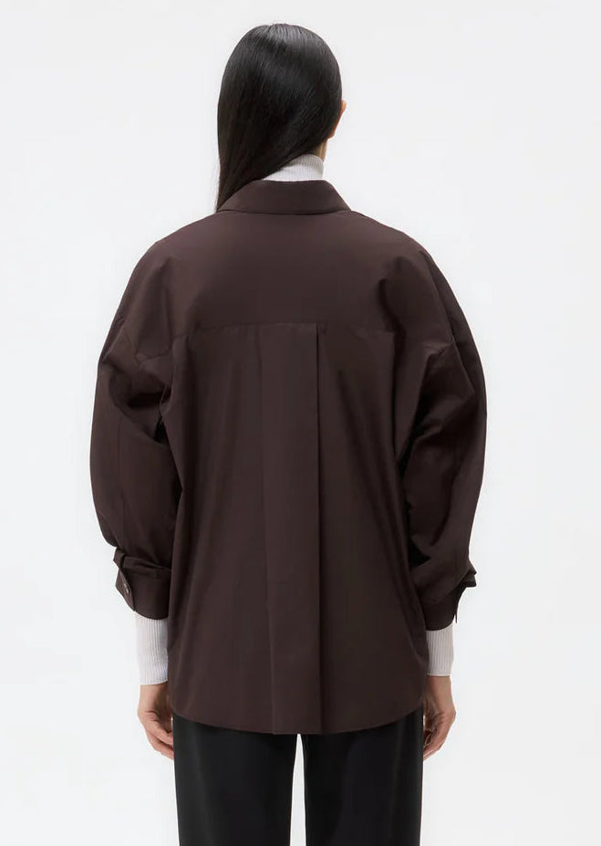 A person with long dark hair is shown from the back against a plain white background, wearing the TWP New Earl Shirt in dark brown over a white turtleneck and black pants.
