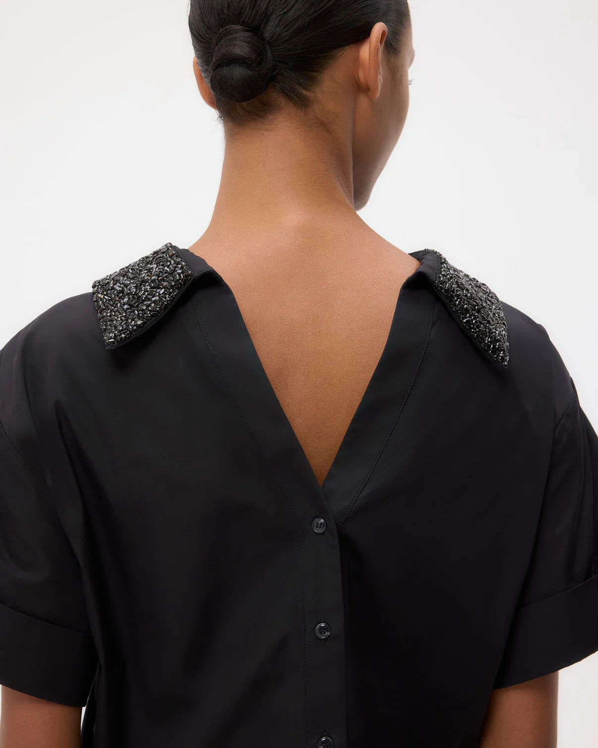 A person with their back to the camera wears the TWP Forever and Always Crystal Collar Shirt, featuring a deep V-shaped back, button details, and embellished shoulders.