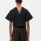 A person stands facing away, wearing the TWP Forever and Always Crystal Collar Shirt with decorative shoulders and a deep V-back, paired with dark pants. Hair is styled in a neat bun.