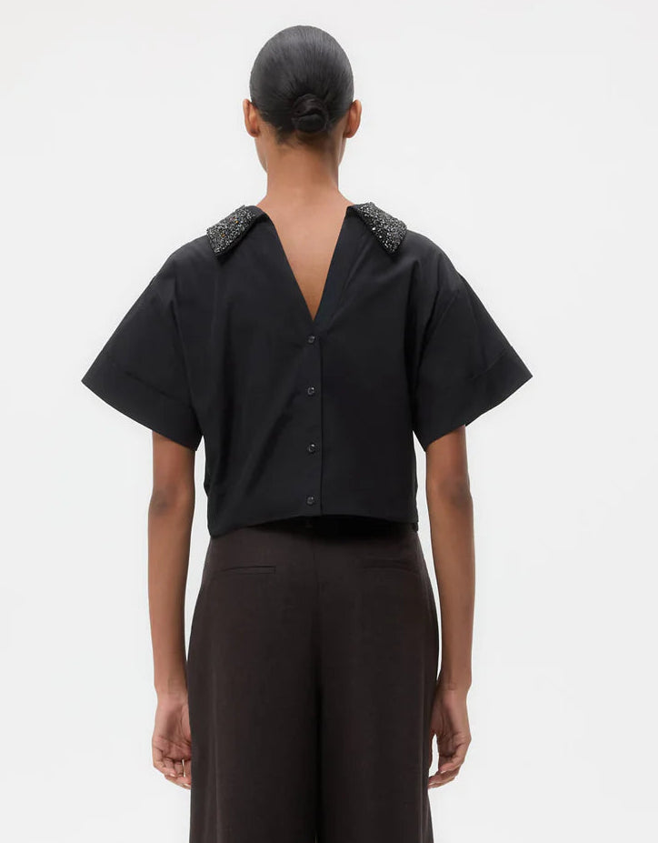 A person stands facing away, wearing the TWP Forever and Always Crystal Collar Shirt with decorative shoulders and a deep V-back, paired with dark pants. Hair is styled in a neat bun.