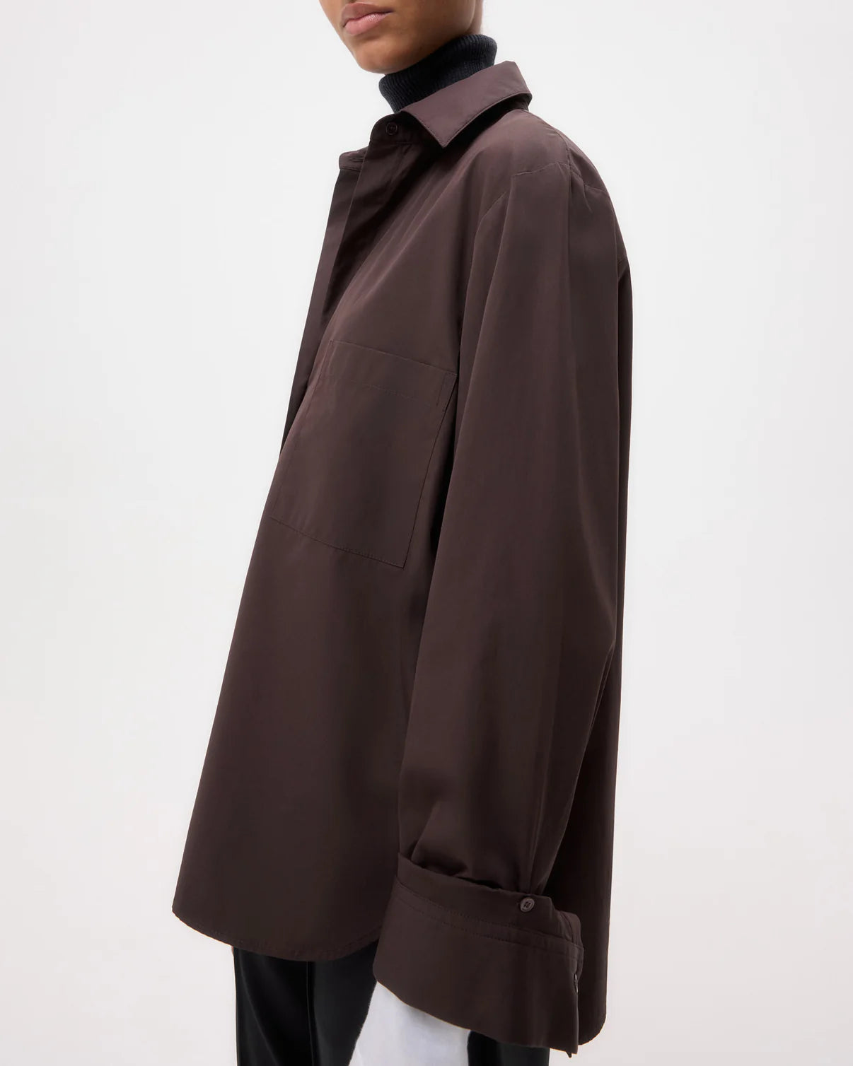 A person wears the TWP Next Morning Shirt, an oversized dark brown button-down with a chest pocket and relaxed fit, layered over a black turtleneck against a plain light background.