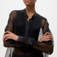 Model wears the TWP Peyton Silk Organza Shirt—an oversized sheer black blouse with pleated front detail—standing with arms crossed against a plain background.