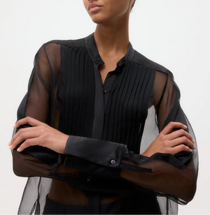 Model wears the TWP Peyton Silk Organza Shirt—an oversized sheer black blouse with pleated front detail—standing with arms crossed against a plain background.