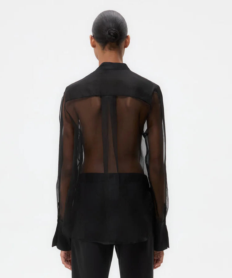 A person faces away, wearing the TWP Peyton Silk Organza Shirt—an oversized sheer design with visible back, opaque cuffs, collar, and lower section. Hair is in a neat bun. Plain white background.