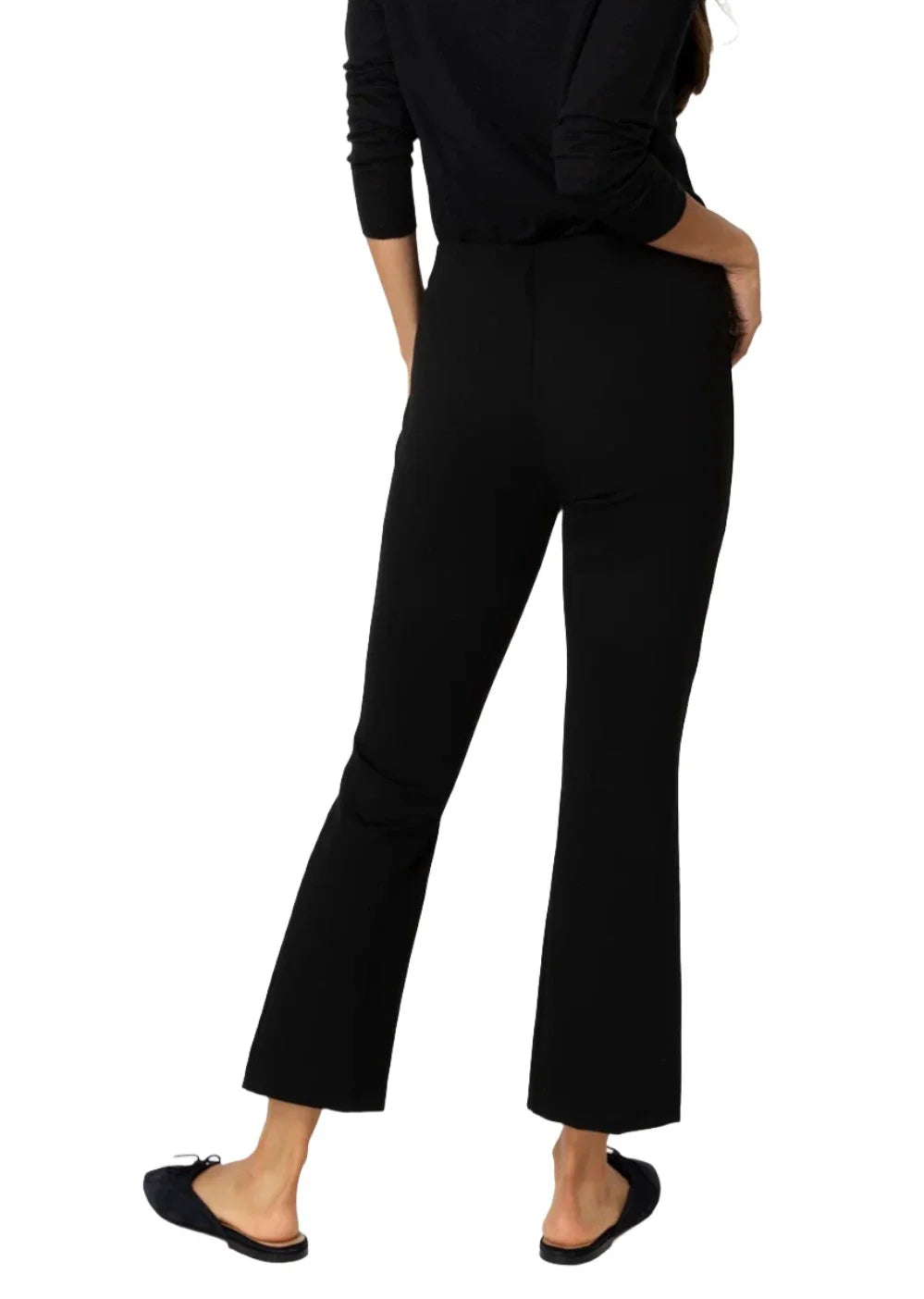 A person wearing a black long-sleeved top, Ann Mashburn Faye Flare Cropped Ponte Knit Pant with an elastic waist, and black shoes stands with their back to the camera, one hand resting on their hip.