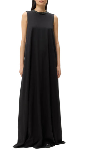 A woman stands against a white background wearing the TWP Capitol Crystal Collar Dress, a sleeveless, floor-length black gown with an A-line silhouette and an embellished crystal collar.