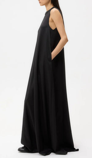 A woman stands sideways against a plain white background wearing the TWP Capitol Crystal Collar Dress, a sleeveless, floor-length black gown with an A-line silhouette and pockets.