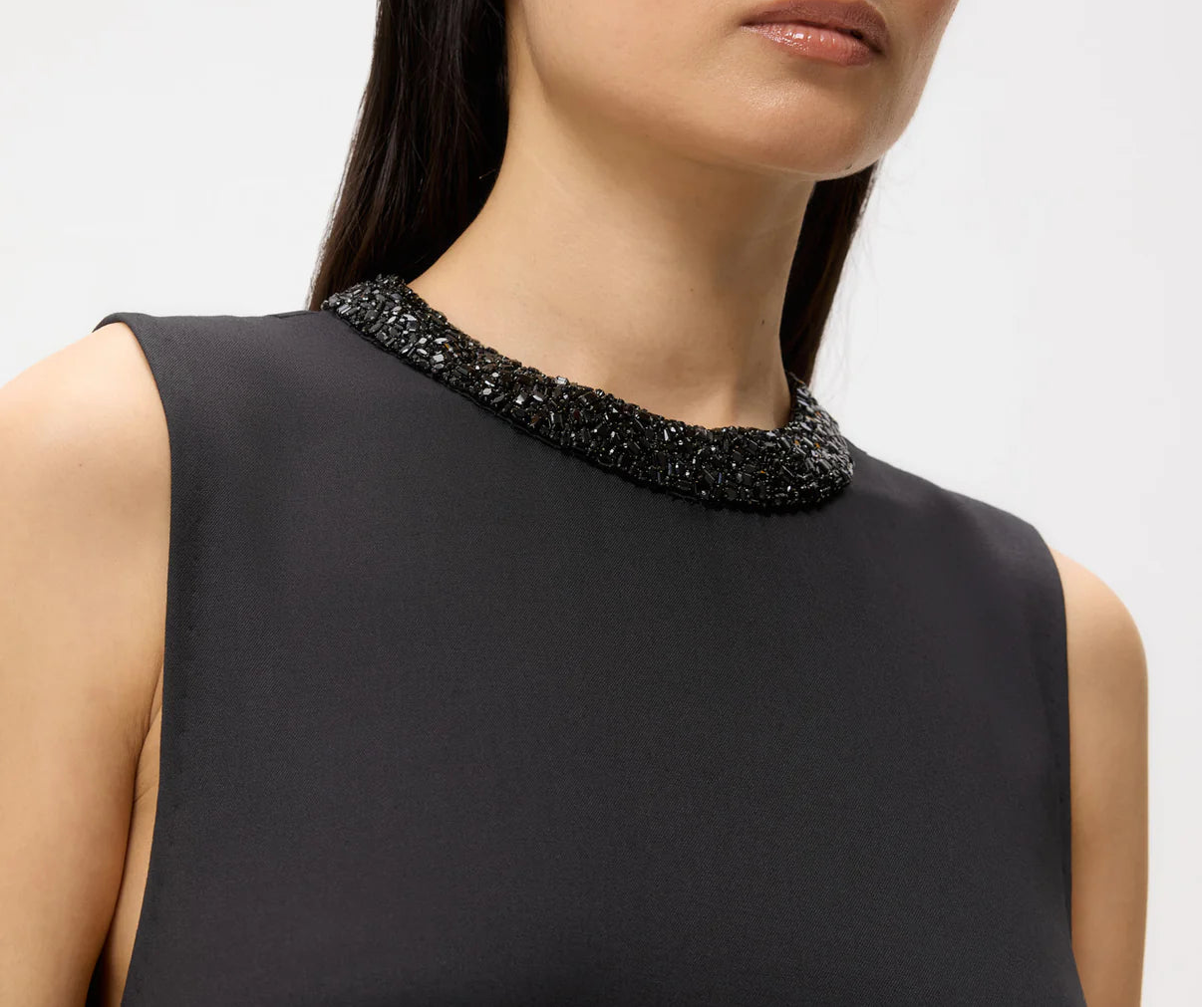 A woman wears the TWP Capitol Crystal Collar Dress, featuring a sleeveless black design and a high crystal collar, shown from the shoulders up against a plain background.