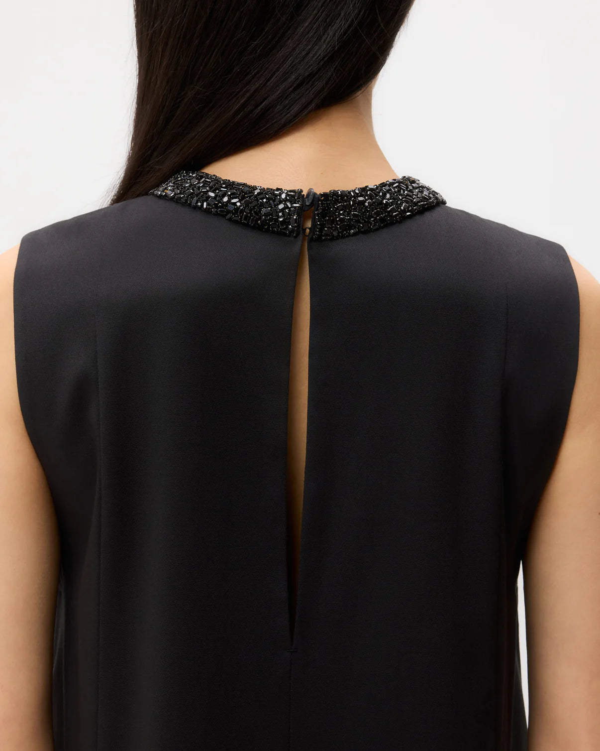 A close-up of a person wearing the TWP Capitol Crystal Collar Dress, a sleeveless black dress featuring a beaded neckline, crystal collar, and keyhole opening at the back.