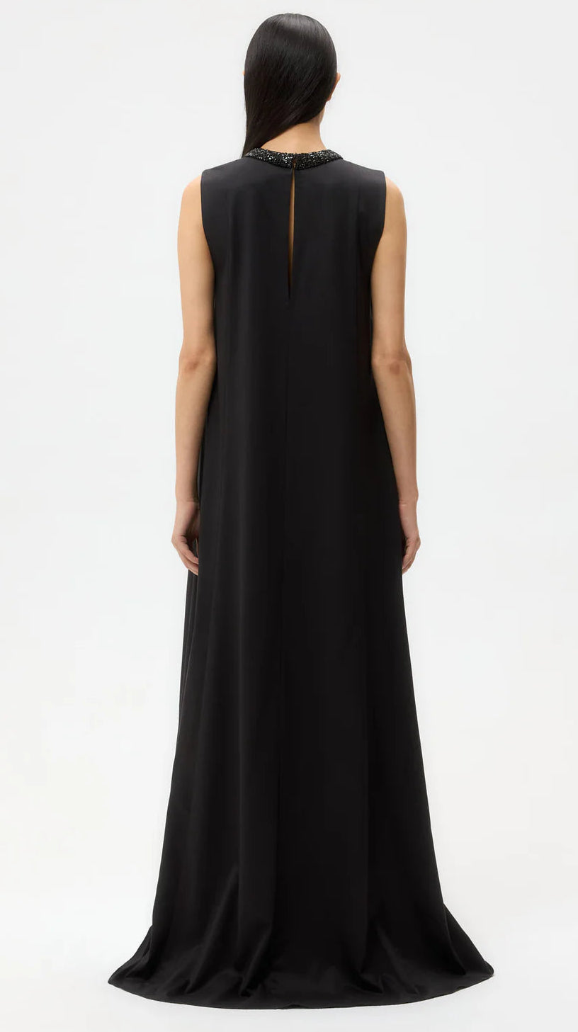 A person stands facing away, wearing the TWP Capitol Crystal Collar Dress—a long, sleeveless silk-finish gown with an A-line silhouette, high neckline, and small back keyhole—set against a plain white background.