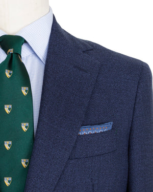 Close-up of a mannequin wearing a Sid Mashburn Kincaid No. 2 Jacket suit jacket with a patterned pocket square, a green tie with shield emblems, and a light blue checked shirt.