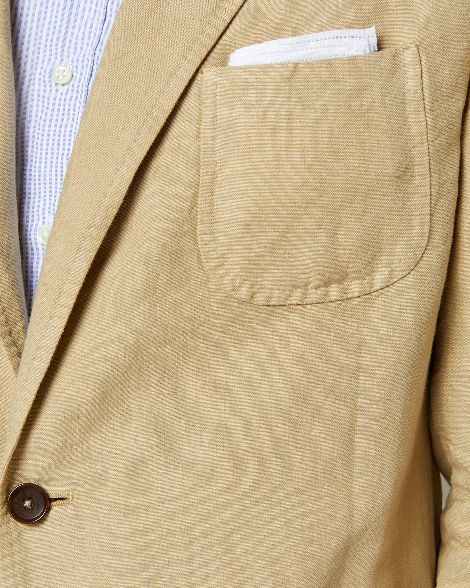 Close-up of a beige blazer made from Italian canvas with a chest pocket and visible stitching. The pocket contains a white handkerchief. The blazer, reminiscent of the Sid Mashburn Butcher Jacket, is worn over a blue and white striped shirt.