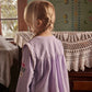 A young child with blond hair in a braid stands indoors, facing away, wearing the Louise Misha Baby Girls’ and Girls’ Anette Dress in light purple corduroy, featuring an embroidered yoke and delicate floral embroidery on the sleeve. Crochet tablecloths cover the furniture.