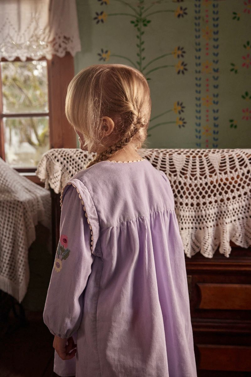 A young child with blond hair in a braid stands indoors, facing away, wearing the Louise Misha Baby Girls’ and Girls’ Anette Dress in light purple corduroy, featuring an embroidered yoke and delicate floral embroidery on the sleeve. Crochet tablecloths cover the furniture.