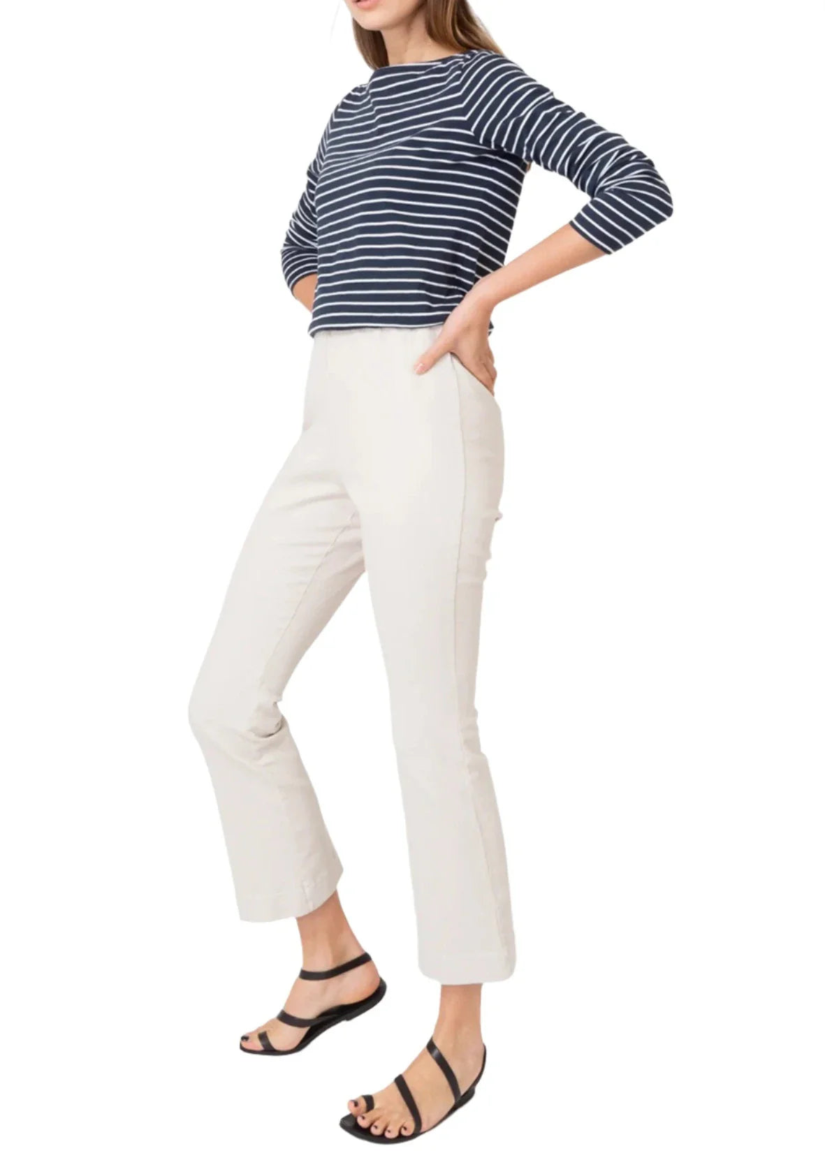 A person wearing a navy and white striped long-sleeve top, Ann Mashburn Faye Flare Cropped Stretch Twill Pant, and black sandals stands with one hand in a pocket against a white background.