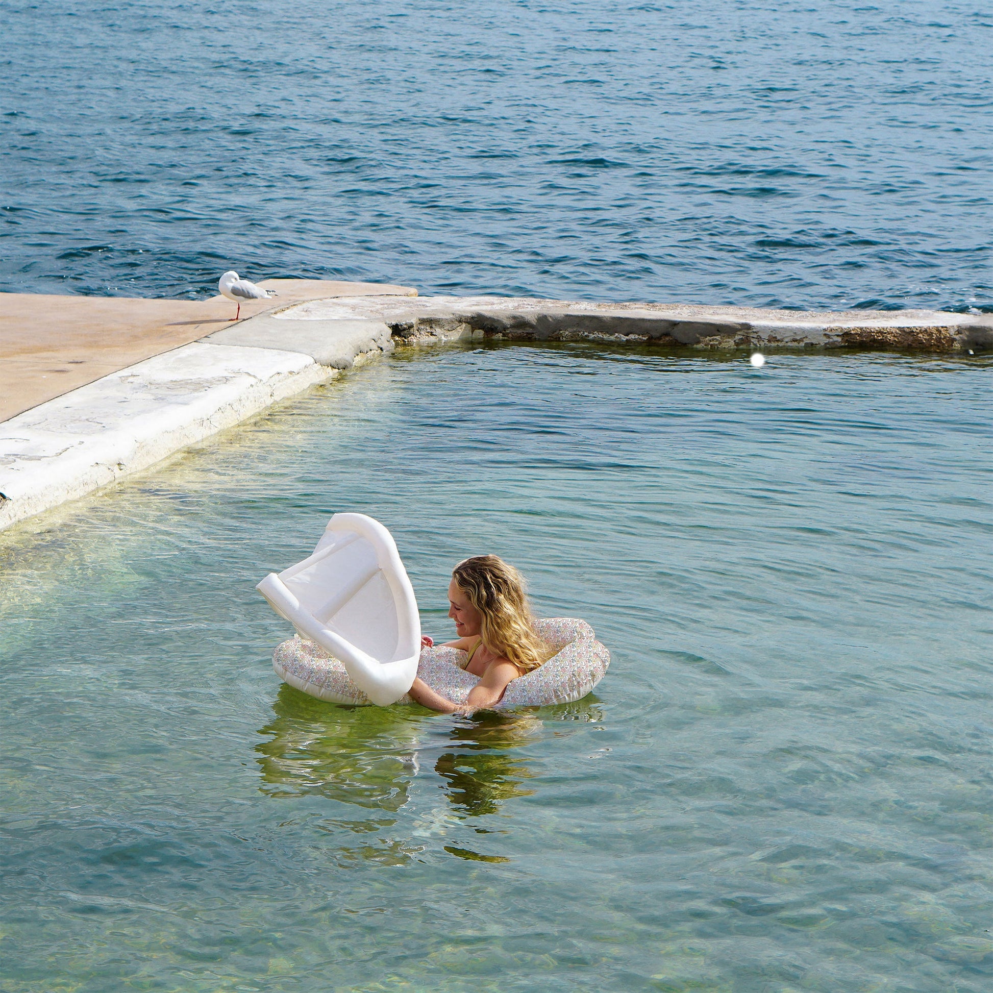A woman sits on the Sunnylife Garden Ditsy Floral Float Together Baby Seat in shallow, clear water near a concrete edge, while a seagull stands on the pavement beside the sea.