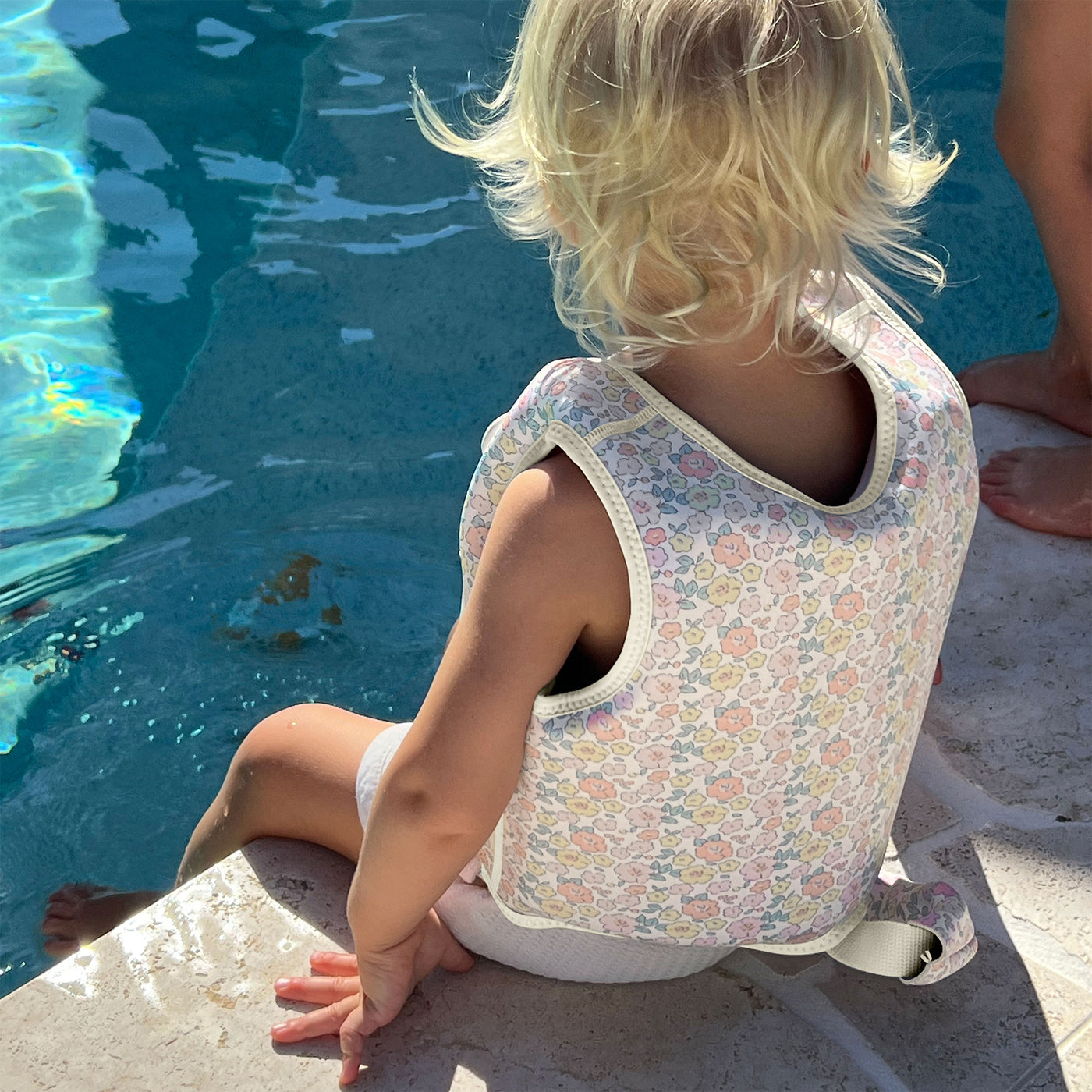 A young child with blonde hair, wearing a Sunnylife Kids' Swim Vest featuring an anti-rise safety strap and a neoprene comfort guard, sits on the edge of a pool with one foot in the water as sunlight shines on them.