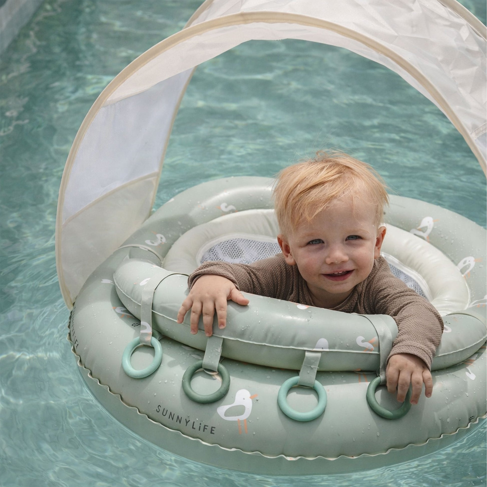 A young child sits in the Sunnylife Sammy Seagull Interactive Baby Float, a light green float with a UPF40 canopy, in a pool, smiling and holding onto the inflatable’s handles.