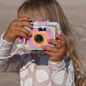 A child with long blonde hair stands outdoors wearing a patterned long-sleeve top, holding the Sunnylife Magenta Underwater Camera up to their face.