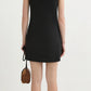 A woman with wavy, shoulder-length hair wears the POSSE Neve Mini Dress—a chic, sleeveless black linen mini dress—and holds a small brown bag, standing with her back to the camera against a white background.