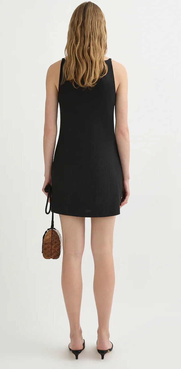 A woman with wavy, shoulder-length hair wears the POSSE Neve Mini Dress—a chic, sleeveless black linen mini dress—and holds a small brown bag, standing with her back to the camera against a white background.