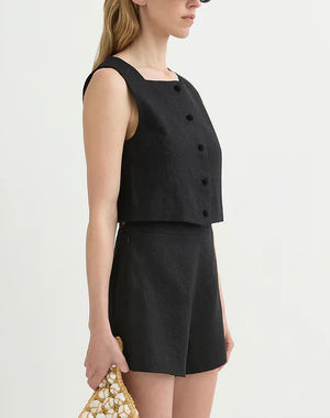 A woman wears the POSSE Rio Top, a sleeveless black buttoned crop top with a high neckline, paired with matching black high-waisted shorts and holds a small beaded handbag.