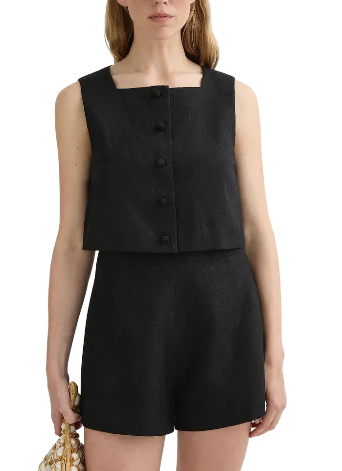 A woman wears the POSSE Rio Top—a black, sleeveless, square-neck button-front top—paired with matching high-waisted mid-weight linen shorts and holds a textured bag in one hand.