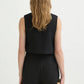 A woman with wavy, shoulder-length blonde hair is seen from behind wearing the POSSE Rio Top—a relaxed fit, sleeveless black top—paired with matching black shorts and holding a small beige woven clutch.