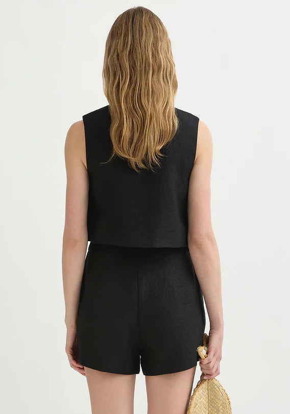 A woman with wavy, shoulder-length blonde hair is seen from behind wearing the POSSE Rio Top—a relaxed fit, sleeveless black top—paired with matching black shorts and holding a small beige woven clutch.