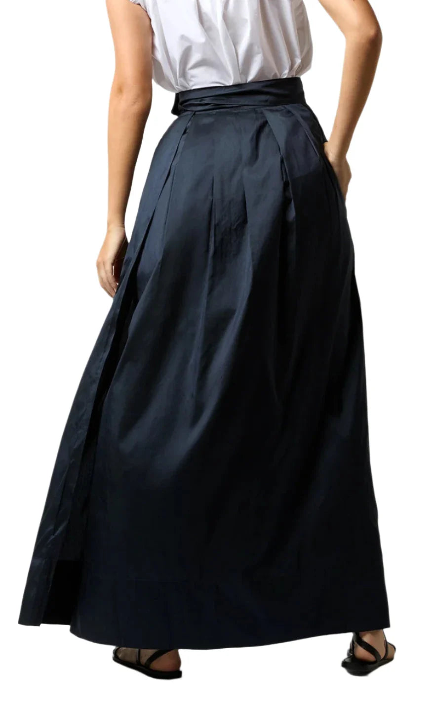 A person wearing a white short-sleeve top and the Ann Mashburn Silk Shantung Pleated Wrap Skirt in dark blue, standing with their back facing the camera.