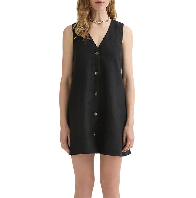 A woman models the POSSE Nancy Mini Dress, a sleeveless black linen mini with a v-neckline, button-front, and straight hem, standing against a white background.