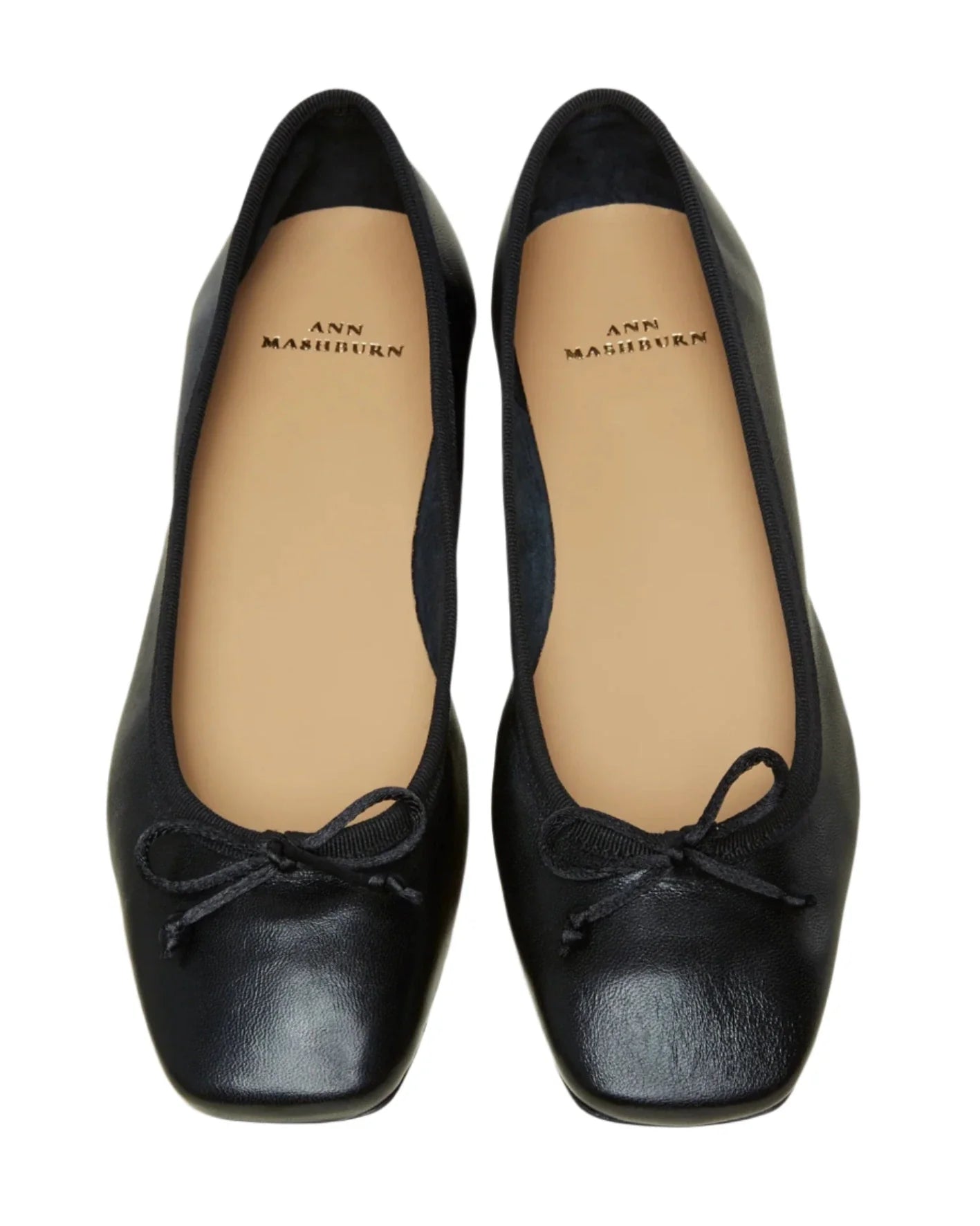 A pair of black Ann Mashburn Square Toe Ballet Flats featuring small bows on the toes, meticulously crafted from luxurious Italian lambskin with cushioned insoles for added comfort and the brand name "Ann Mashburn" printed on them.