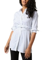 A person wearing the Ann Mashburn Gabrielle Tie Tunic, a white and blue striped tunic made of 100% cotton Italian poplin with long sleeves and a tied belt at the waist, paired with black pants.