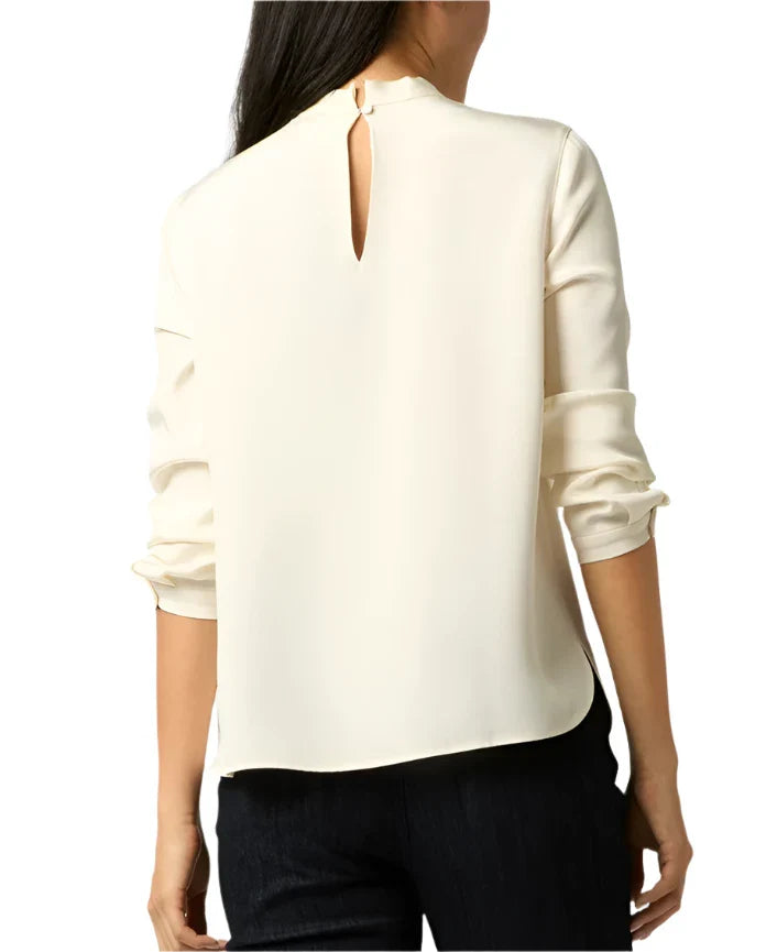 A woman with long dark hair wears the Ann Mashburn Pippa Top, a cream-colored long-sleeve tie-neck blouse with a keyhole back cutout. She stands with her back turned to the camera, showcasing the top’s elegant design.