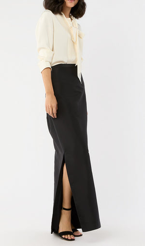 A woman wearing the Ann Mashburn Long Pencil Silk Faille Skirt with a cream silk blouse and black heeled sandals stands against a plain white background.
