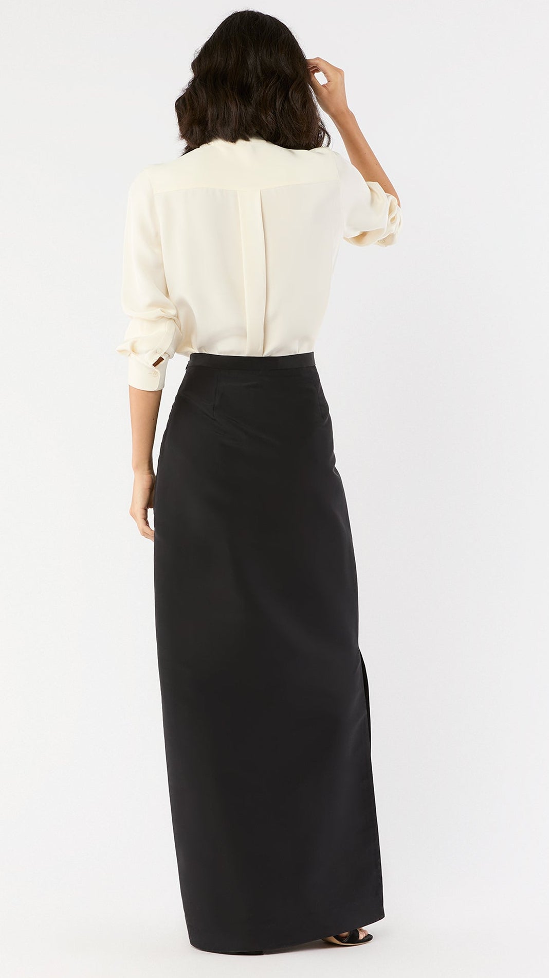 A woman stands facing away, wearing the Ann Mashburn Long Pencil Silk Faille Skirt with a white silk long-sleeve blouse with rolled-up sleeves.