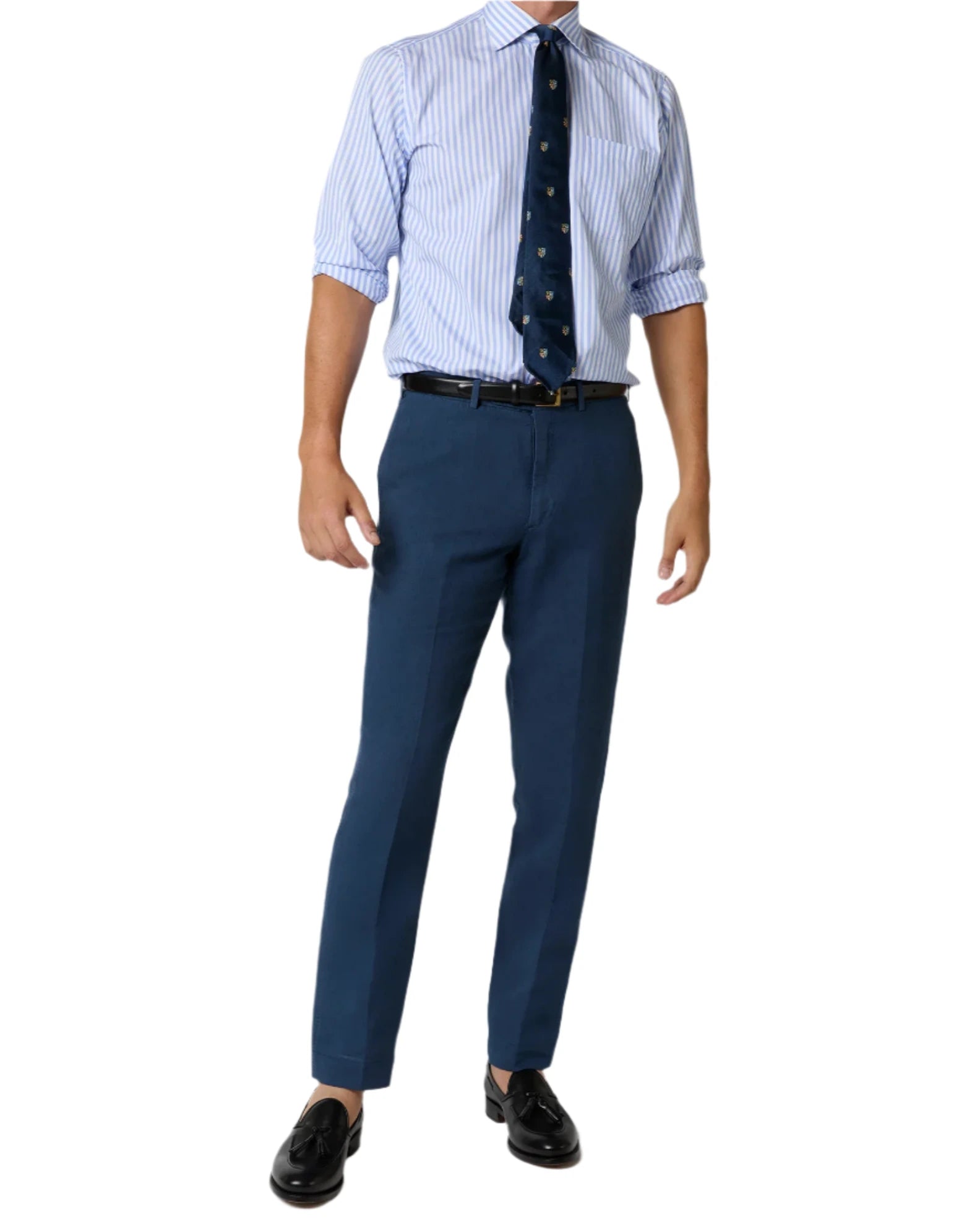 A person in Sid Mashburn Sport Trousers, a light blue striped dress shirt with rolled sleeves, dark blue tie, black belt, and black loafers.