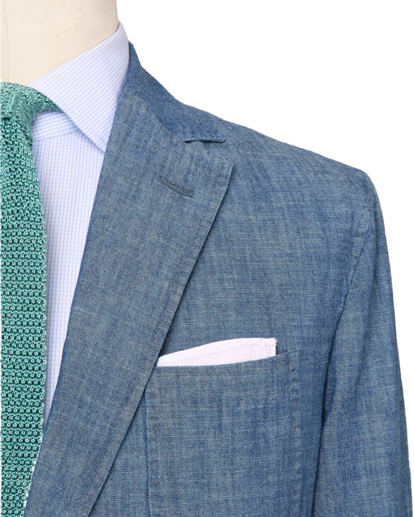 A close-up of a person wearing the Sid Mashburn Butcher Jacket, a blue denim suit jacket with spalla camicia construction, paired with a white pocket square, light blue dress shirt, and green patterned tie.