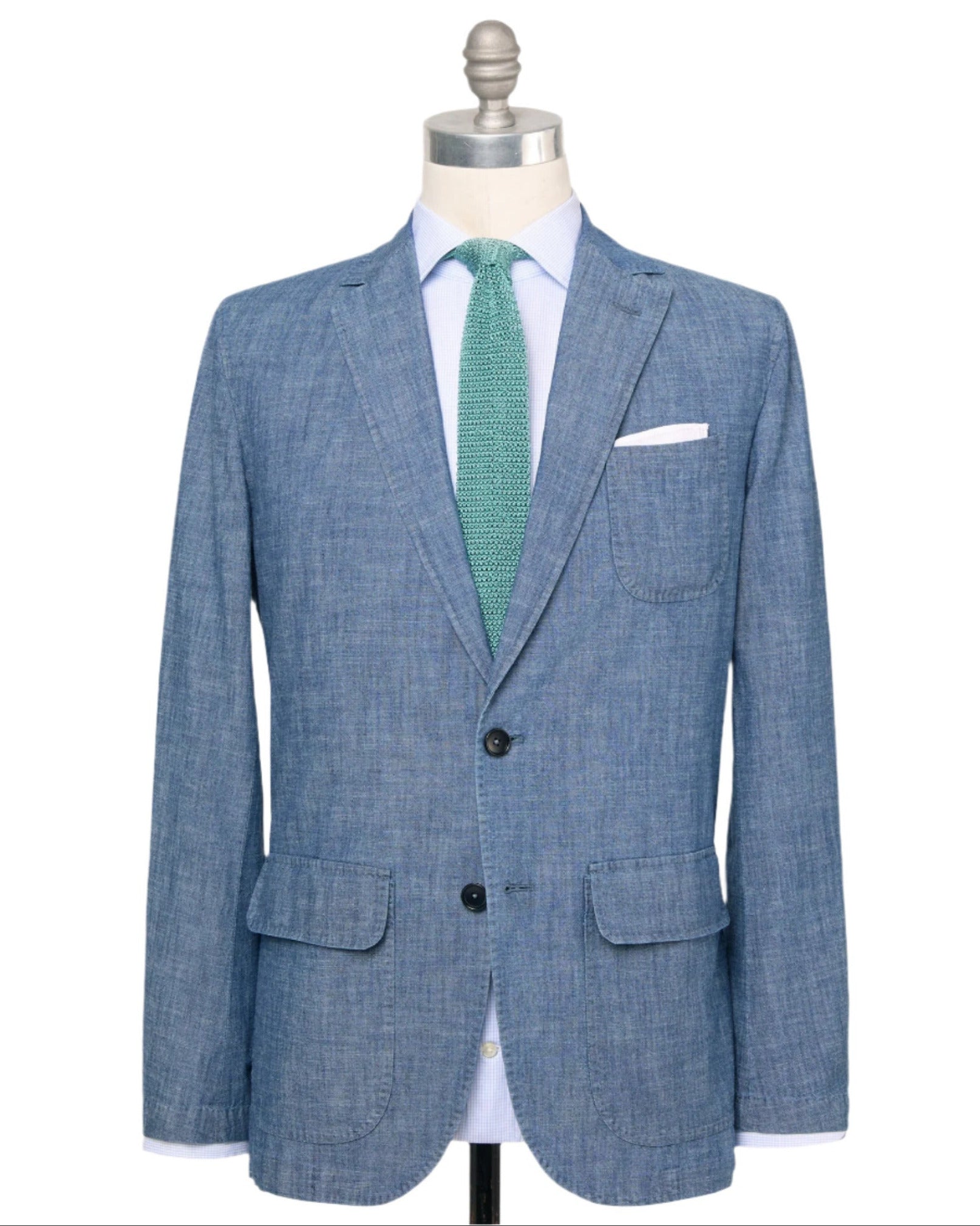The Sid Mashburn Butcher Jacket, a blue Portuguese chambray blazer with notch lapels, spalla camicia construction, two buttons, patch pockets, and a breast pocket, is shown on a mannequin with a white shirt and green textured tie.