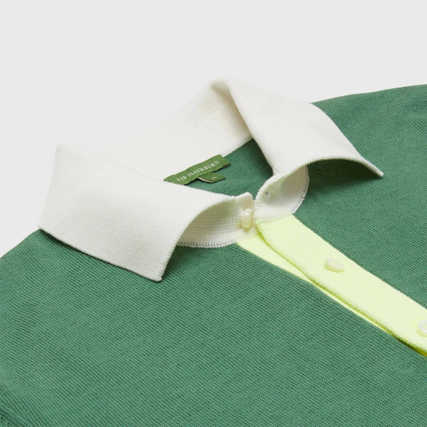 The Sid Mashburn Rally Polo Sweater is a green sweater-knit polo with a white collar and light yellow button placket, made from high-twist cotton and displayed flat on a light background.