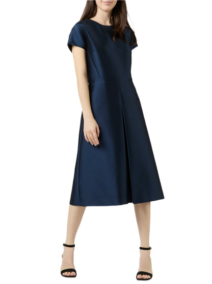 A woman wears the Ann Mashburn Suzzette Dress, a navy blue knee-length pleated A-line with cap sleeves, paired with black high-heeled sandals, standing against a white background.