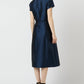 A woman faces away in a knee-length Ann Mashburn Suzzette Dress with an A-line skirt and cap sleeves, styled with black heeled sandals against a plain light background.