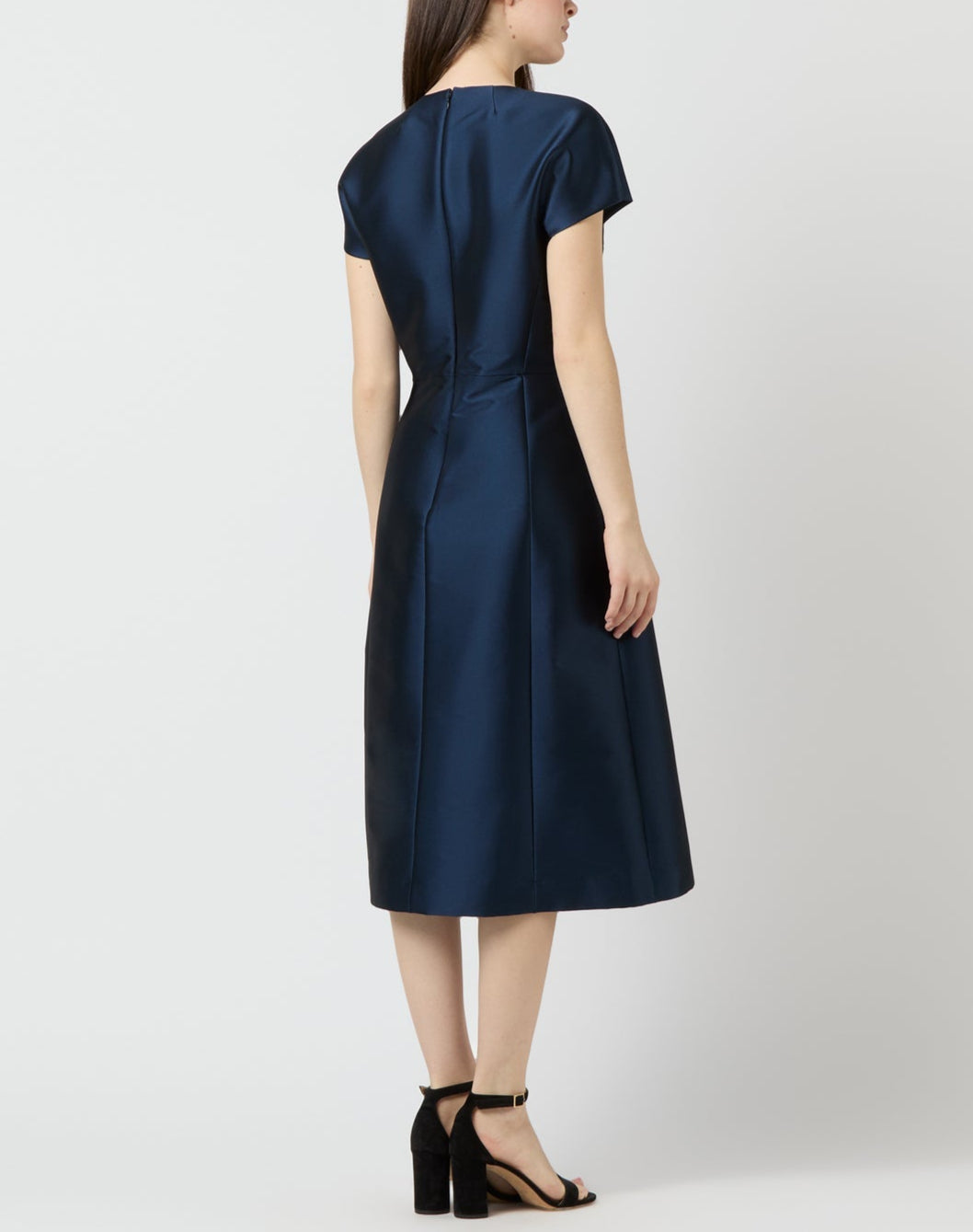 A woman faces away in a knee-length Ann Mashburn Suzzette Dress with an A-line skirt and cap sleeves, styled with black heeled sandals against a plain light background.