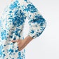 A woman models the Ann Mashburn Francine Dress, featuring white fabric with blue florals and chic bracelet sleeves, standing with one hand in a side pocket against a plain background.