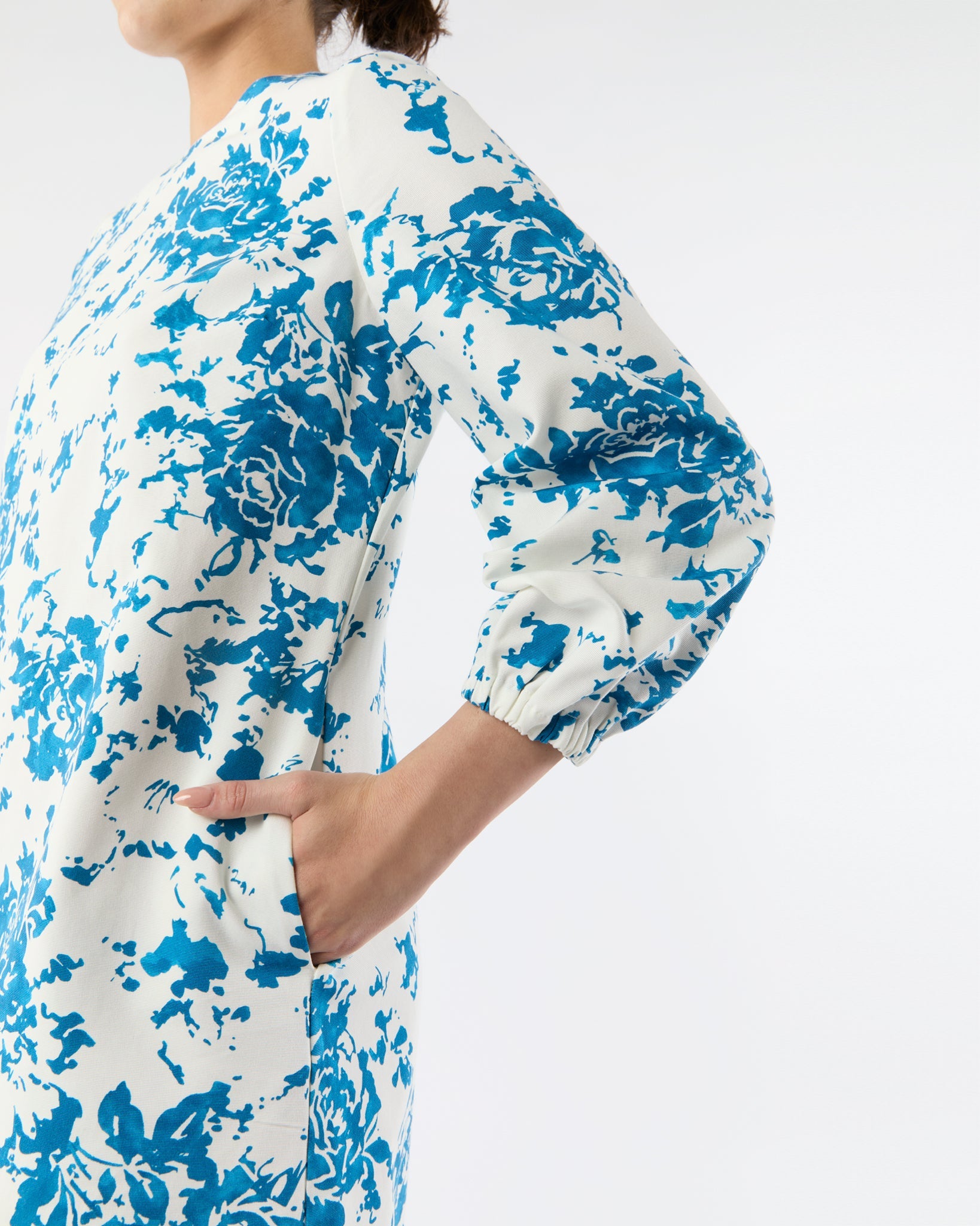 A woman models the Ann Mashburn Francine Dress, featuring white fabric with blue florals and chic bracelet sleeves, standing with one hand in a side pocket against a plain background.